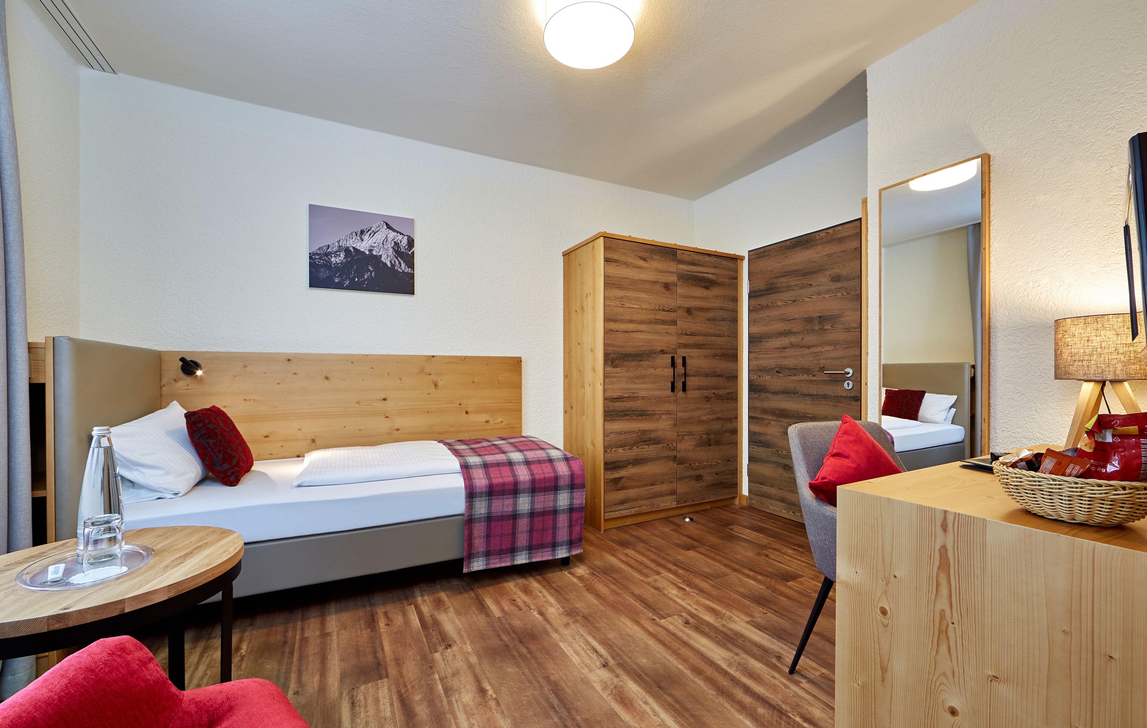 Ein modernes Hotelzimmer mit einem hölzernen Bett, einem kleinen Tisch mit einem Glas, einem Sessel, einem Kleiderschrank und einem Spiegel.