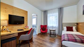 Hotelkamer met een houten bureau, een televisie, een rode stoel, een bed met een ruitendeken en uitzicht op het omliggende landschap door een raam.