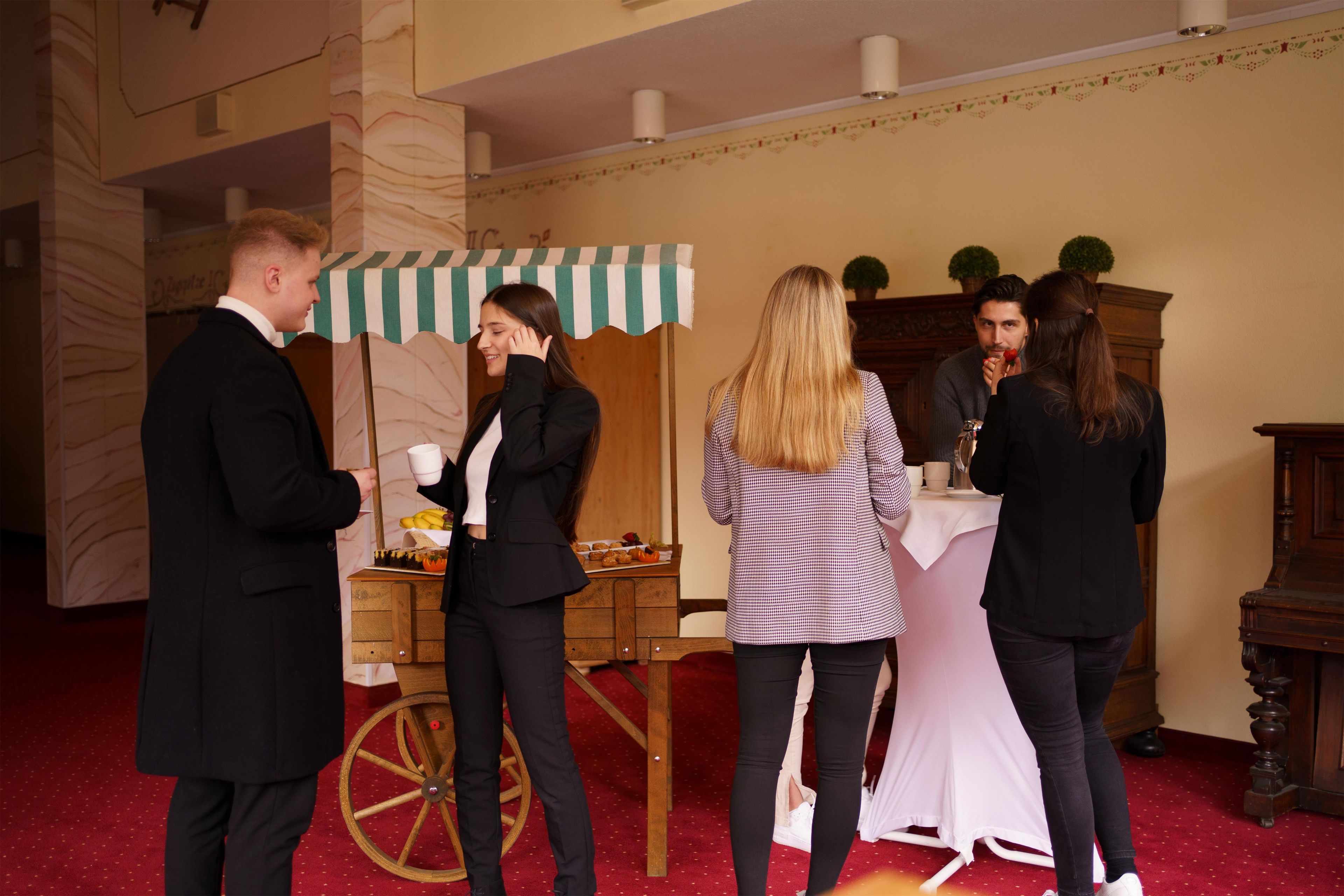 A group of people standing in a room with a red carpet, a wooden cart with food, and a table with a white tablecloth.