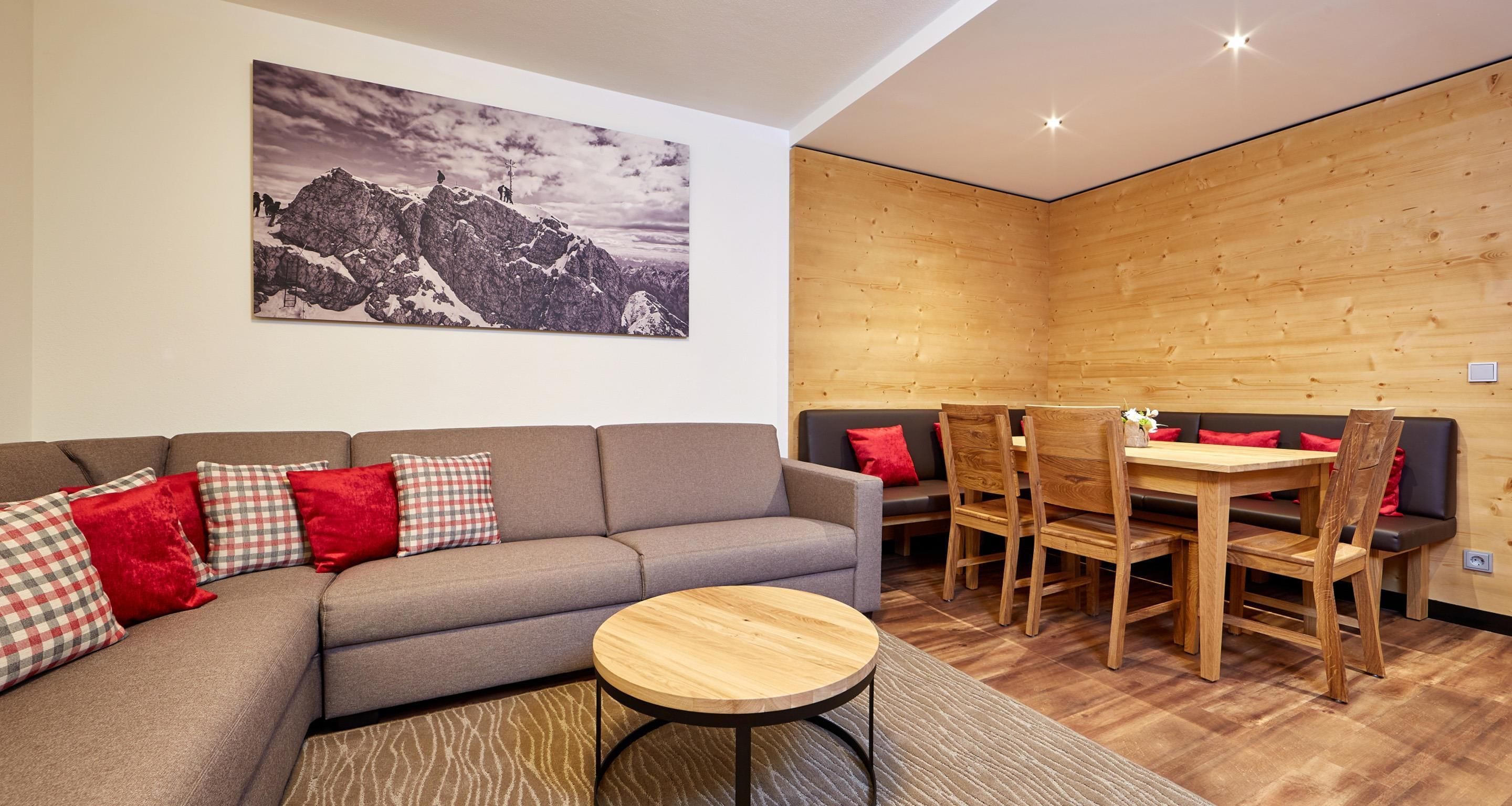 A modern living room with a grey sofa, red and checkered cushions, a round wooden coffee table, and a dining area with wooden chairs and a table.