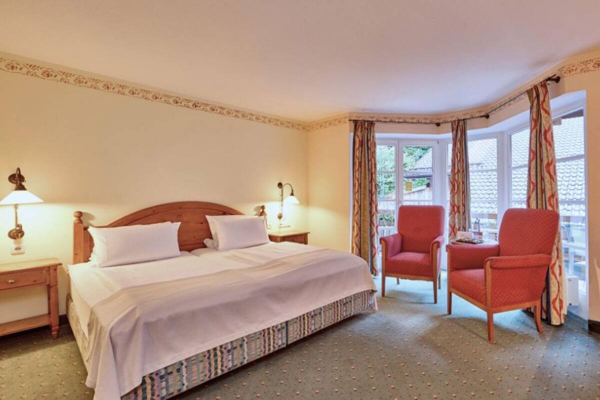 Quarto de hotel com uma cama, duas mesas de cabeceira, uma cadeira de madeira e uma janela com cortinas.