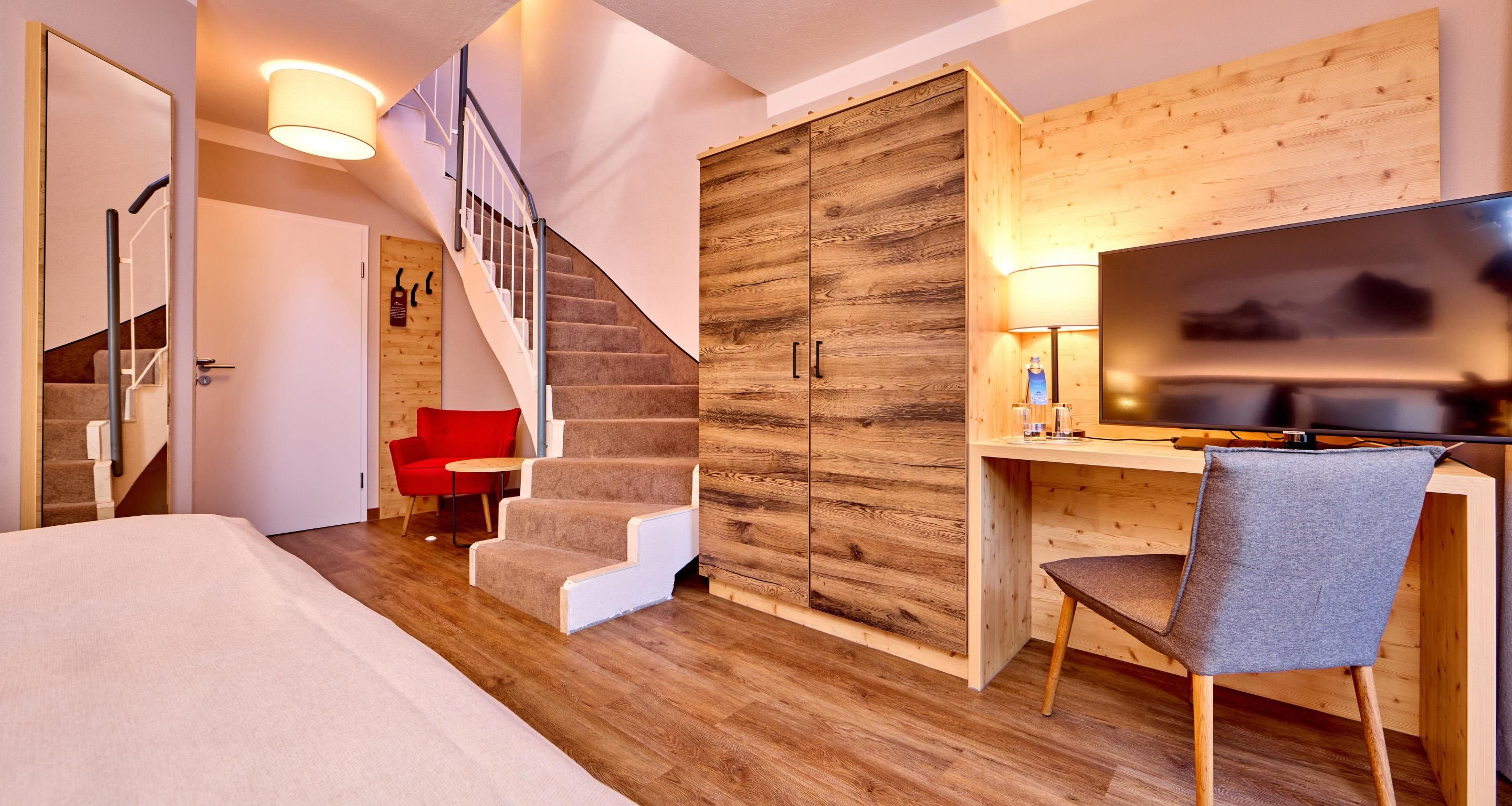 A modern hotel room with a wooden desk, a TV, a staircase, and a red chair.