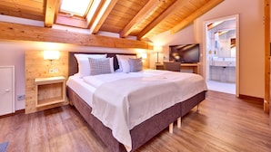 Habitación de hotel moderna con techo de madera, una cama grande con cabecera gris, lámparas de noche, un televisor de pantalla plana y un baño visible a través de una puerta abierta.