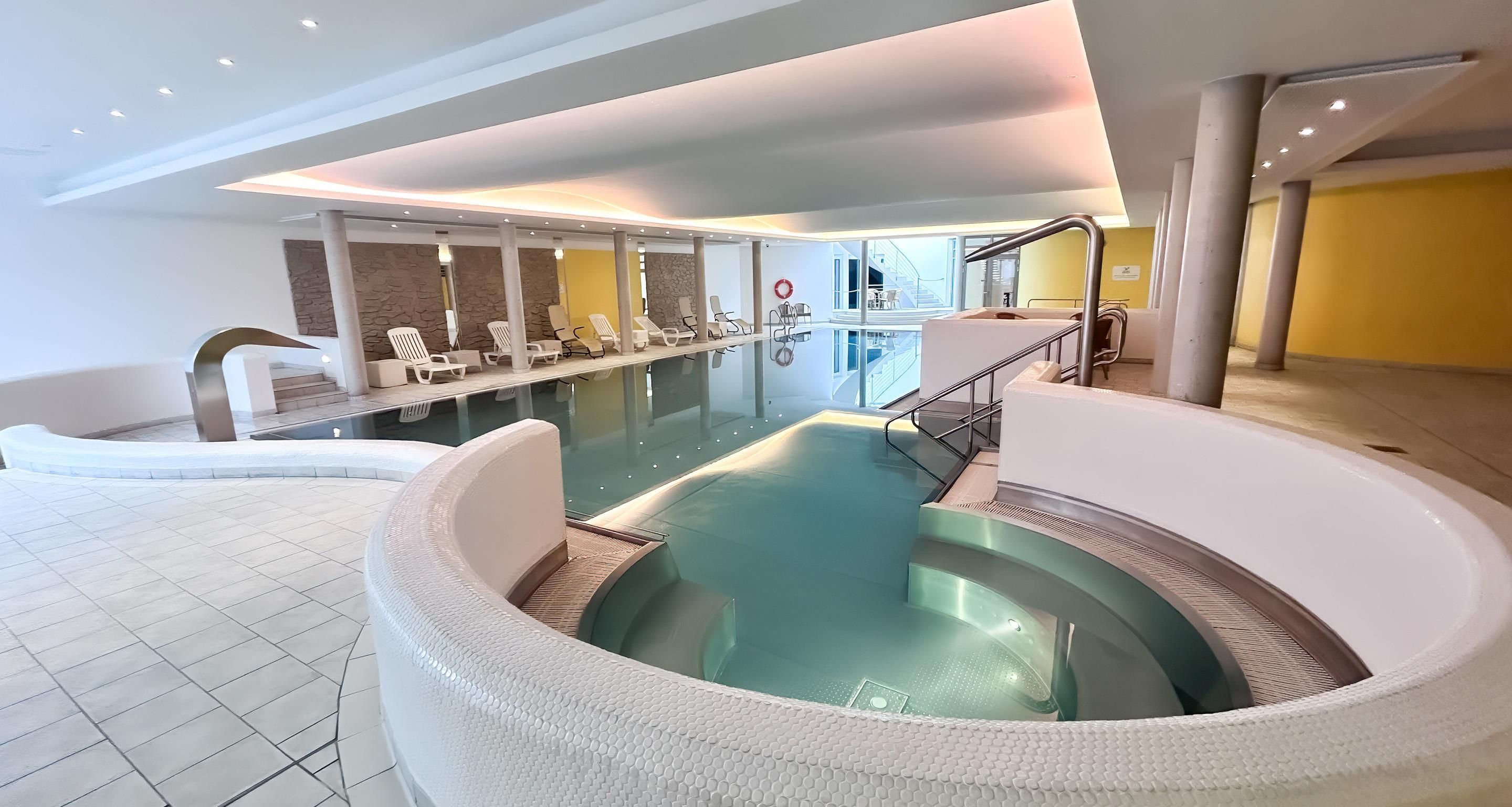 A modern indoor pool area with a hot tub, lounge chairs, and a stone wall.
