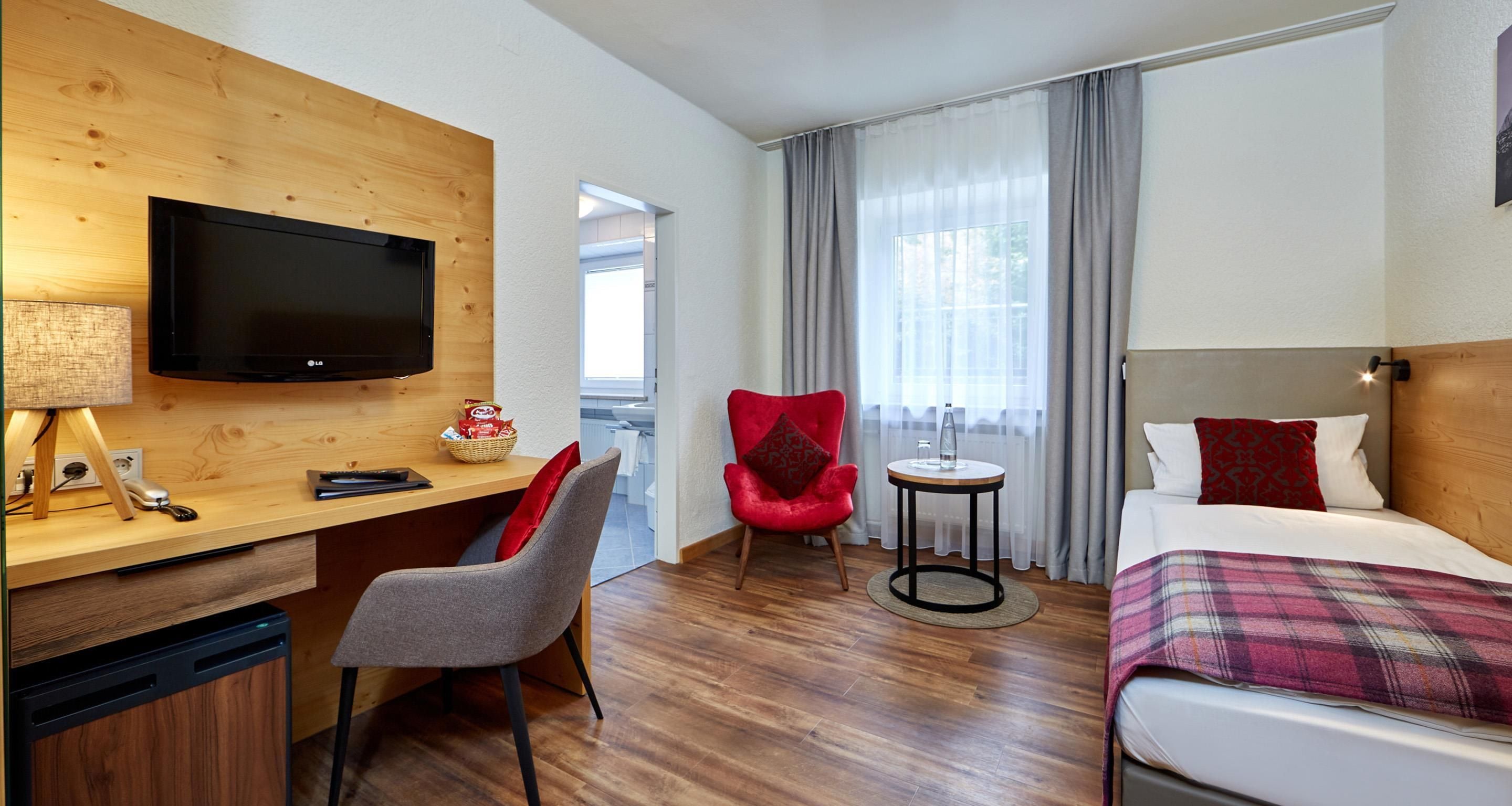 A hotel room with a wooden desk, a television, a red chair, a bed with a plaid blanket, and a window with curtains.