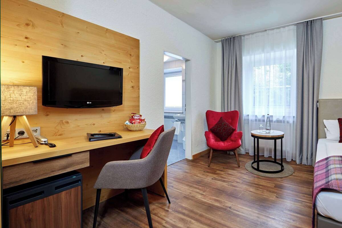 Quarto de hotel com secretária de madeira, televisão, cadeira vermelha, cama com manta xadrez e janela com cortinas.