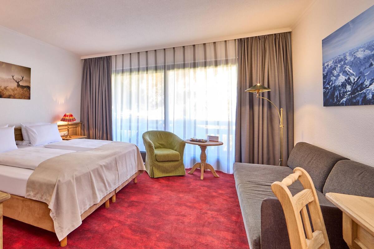 Quarto de hotel com uma cama grande, uma poltrona verde, um sofá cinzento e uma pequena mesa.