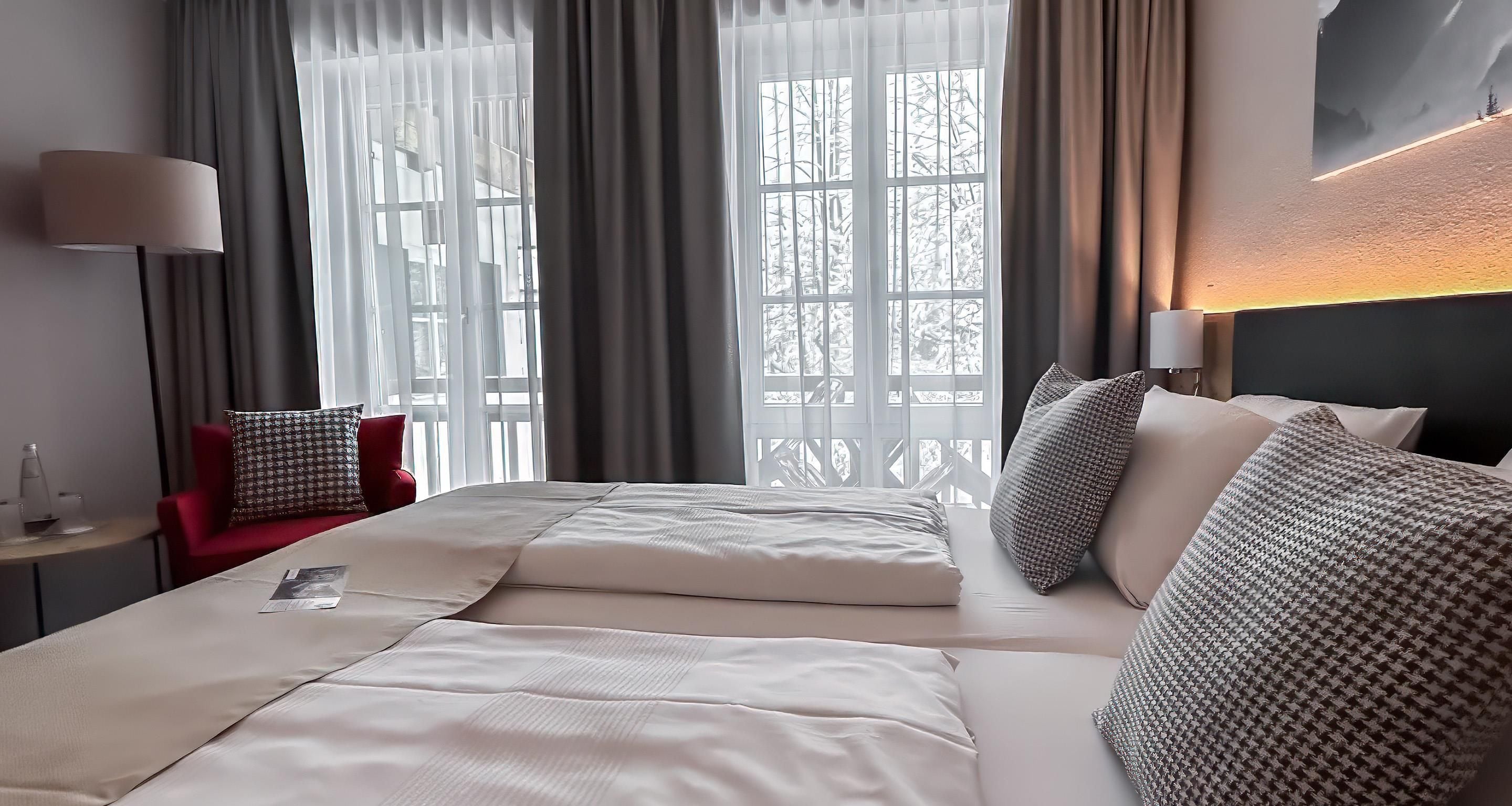 Una habitación de hotel con una cama grande, una mesita de noche, una silla y un ventanal con cortinas.