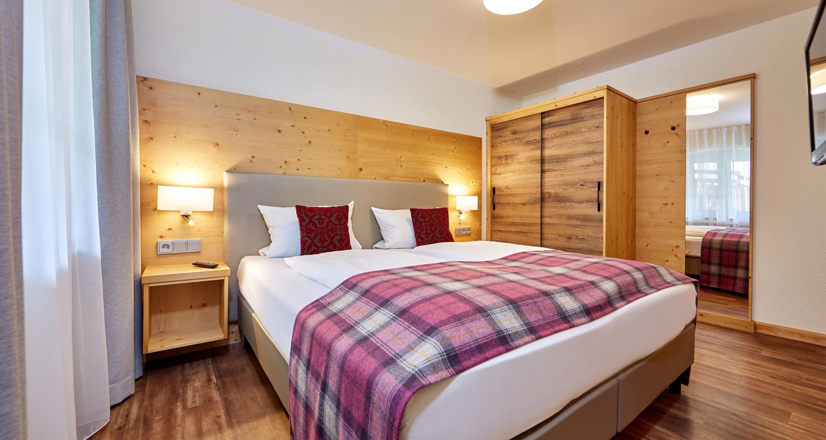 Habitación de hotel con cabecera de madera, cama con manta escocesa, lámparas de noche y un armario con espejo.