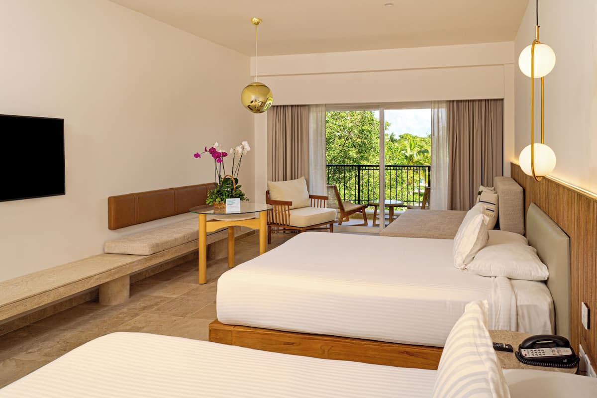 A hotel room with two beds, a TV, a desk with a chair, a balcony with a view of greenery, and a hanging light fixture.