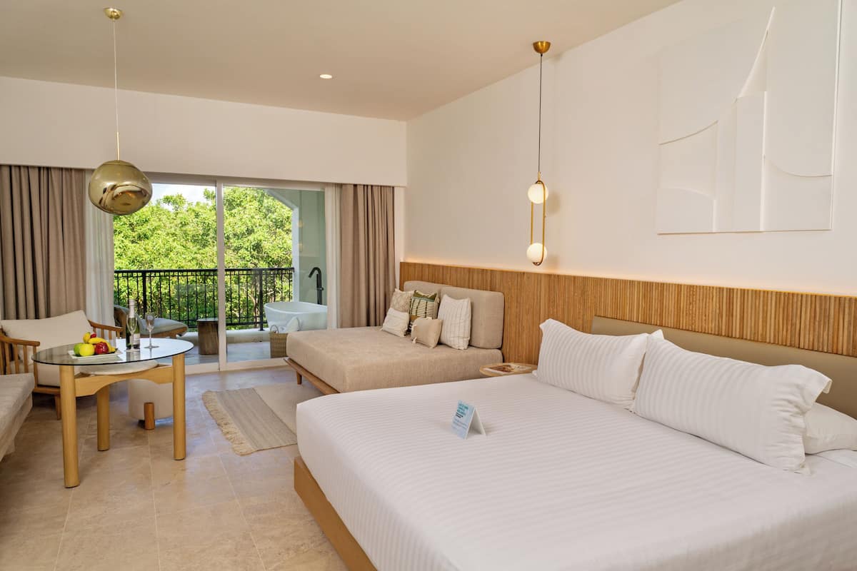 A hotel room with a bed, a desk with a fruit bowl, a chair, a balcony with a view of greenery, and a hanging light fixture.