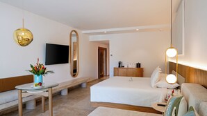 A modern hotel room with a bed, bedside table, a small round table with a vase of flowers, a large mirror, and a flat-screen TV mounted on the wall. - Kimpton Tres Rios All Inclusive by IHG (Playa del Carmen)