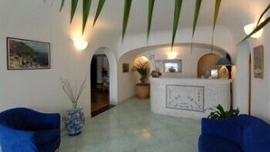 A reception area with a marble reception desk, two blue armchairs, a potted plant, and wall-mounted lighting. - Hotel Le Fioriere (Praiano)