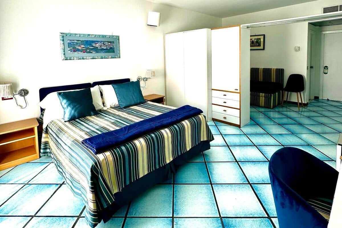 A bedroom with a bed, a chair, a nightstand, a wall-mounted lamp, and a door leading to another room.