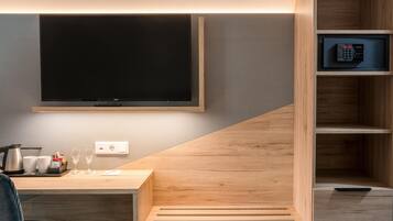 A modern hotel room with a flat-screen TV mounted on the wall, a wooden desk with a kettle, cups, and a remote control.