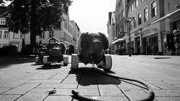 Une photographie en noir et blanc d’une rue, avec une grande statue ornée au premier plan, un arbre à gauche et des bâtiments de chaque côté.