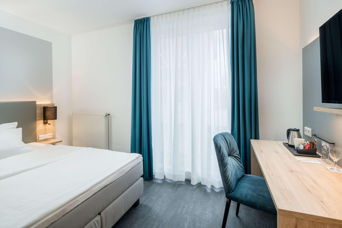 A hotel room with a bed, a desk with a chair, a television, and a window with curtains.