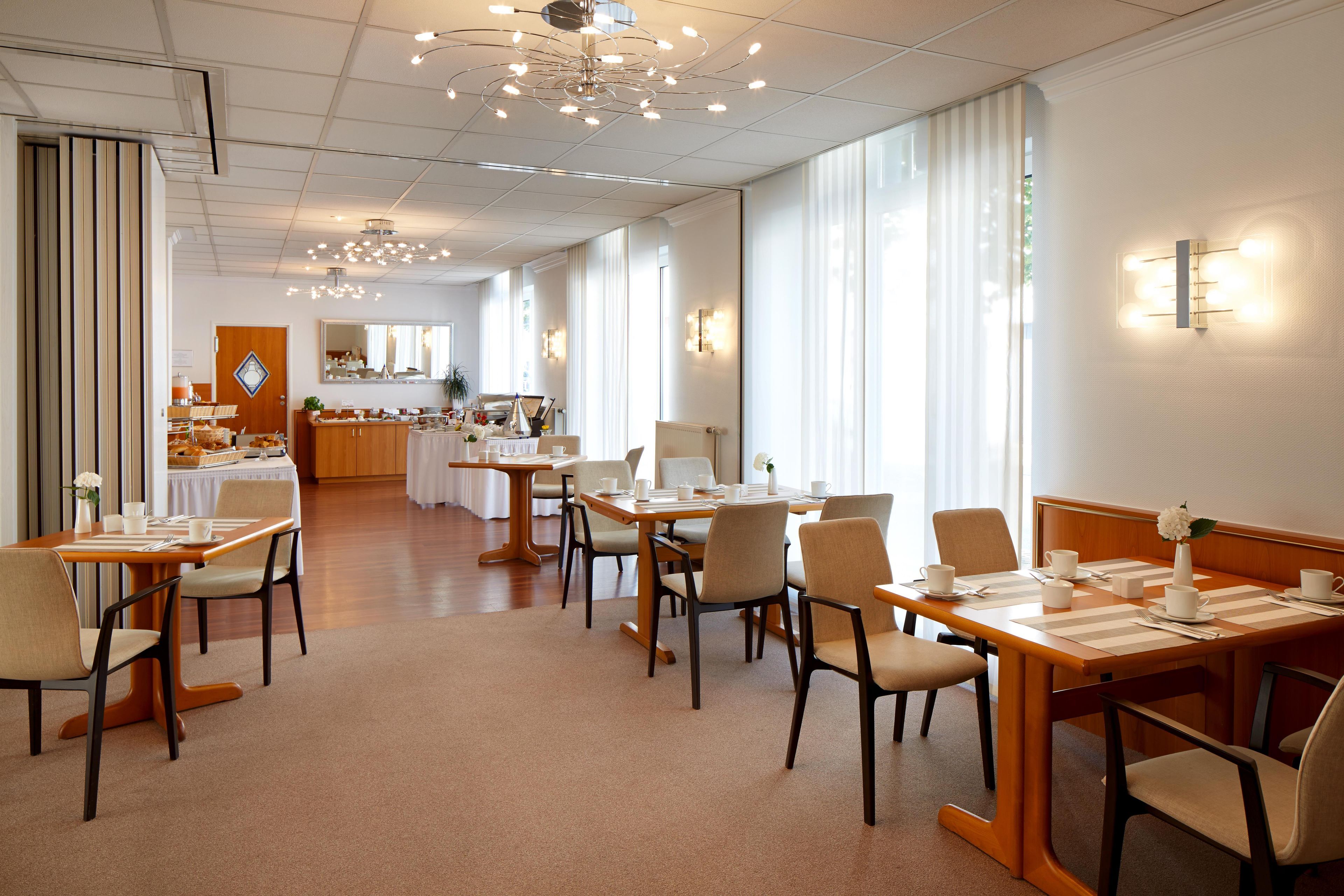 A hotel dining area with multiple tables and chairs, a central chandelier, and a view of the kitchen in the background.