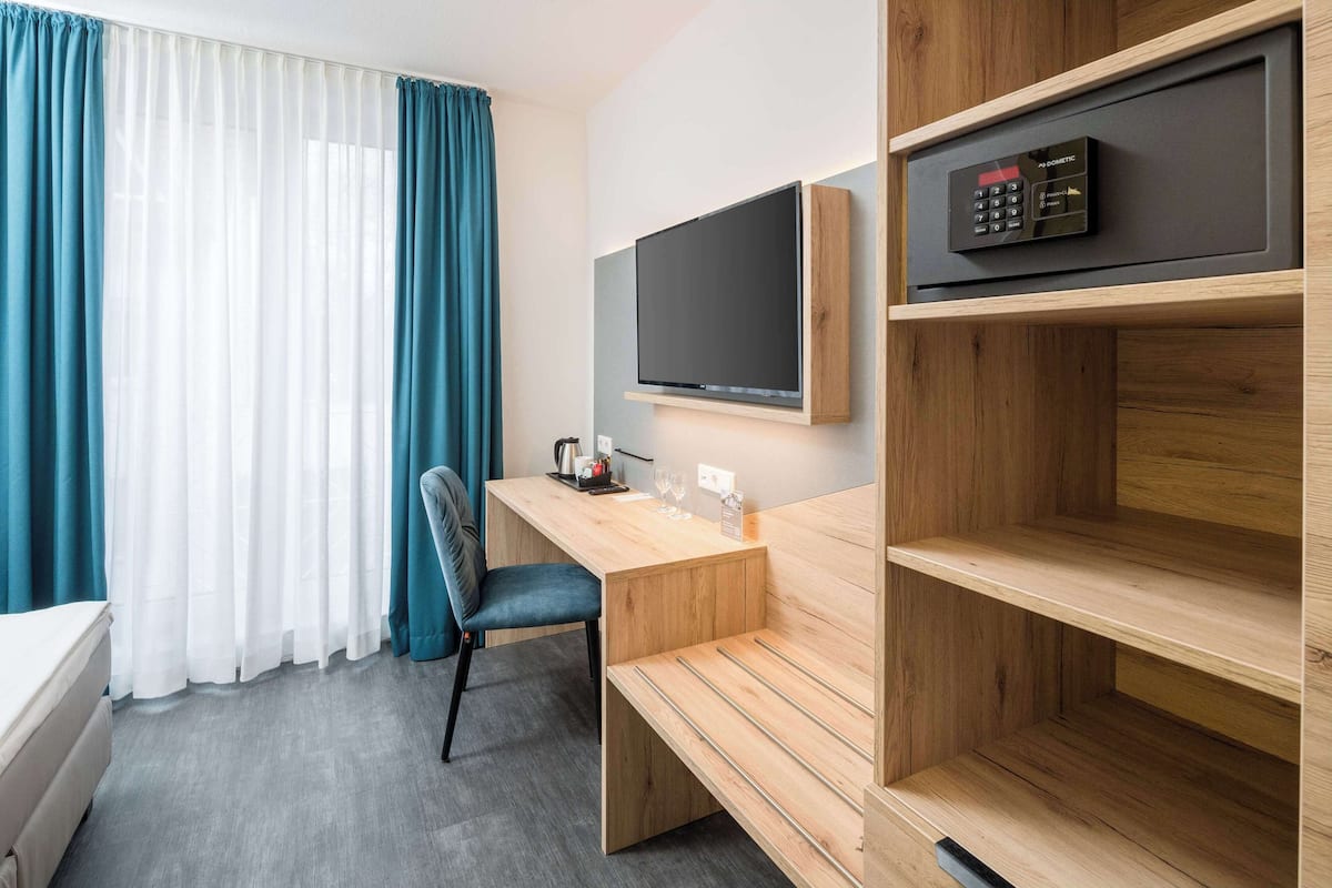 A hotel room with a wooden desk, a TV mounted on the wall, a bed, and a window with curtains.