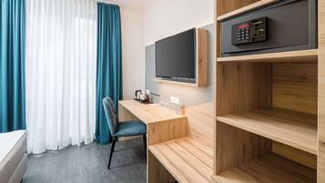 A hotel room with a wooden desk, a TV mounted on the wall, a bed, and a window with curtains.