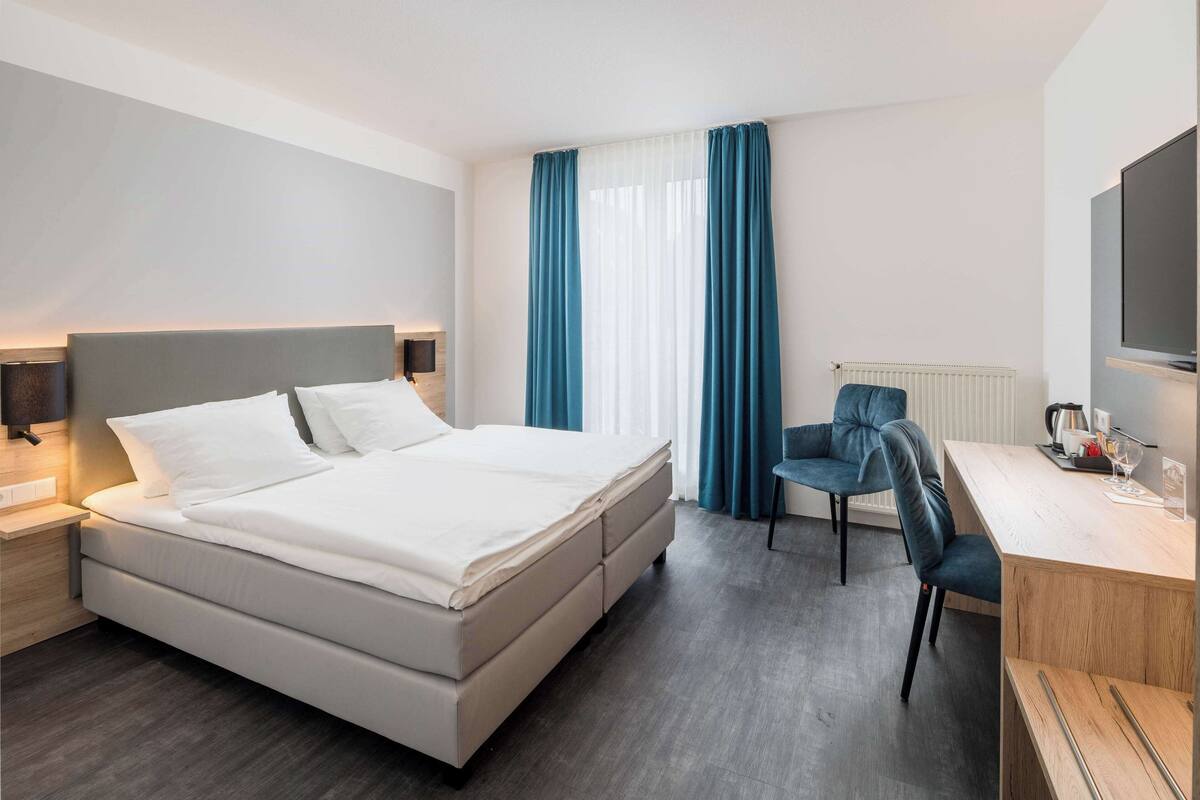 A modern hotel room with a large bed, a desk with a chair, a television, and a window with curtains.