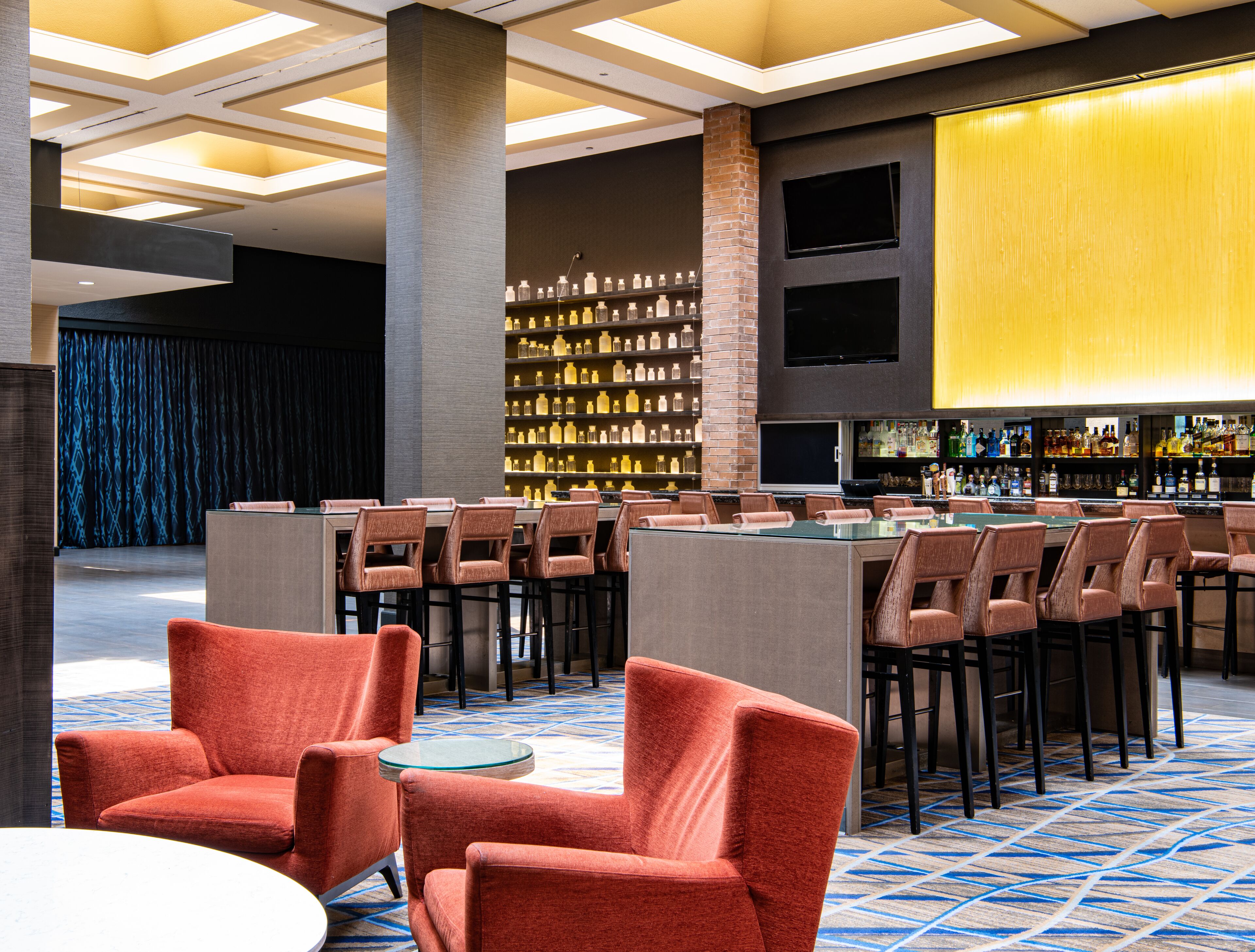 A modern bar area with a long counter, multiple bar stools, and a wall of liquor bottles.