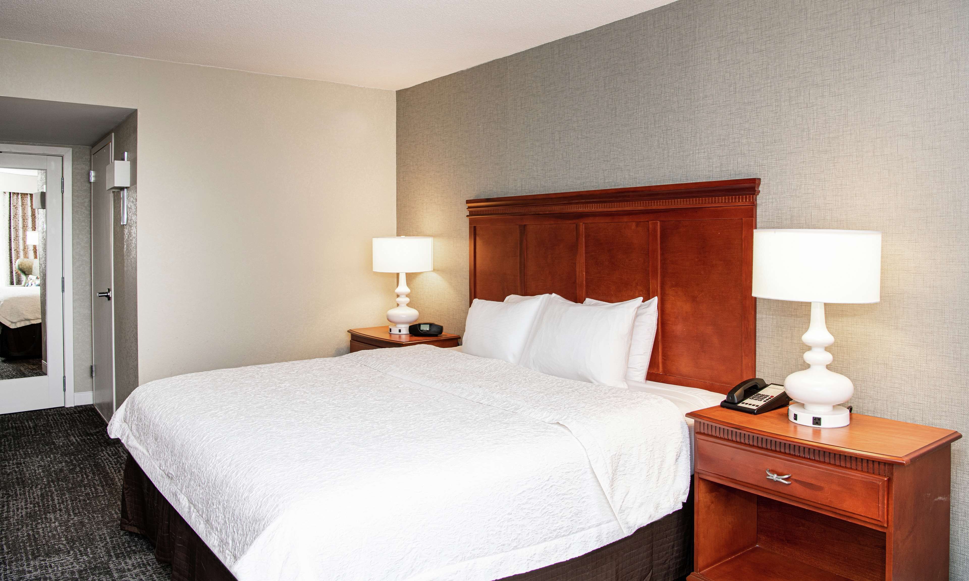 A hotel room with a large bed, two bedside tables with lamps, a wooden headboard, and a carpeted floor.