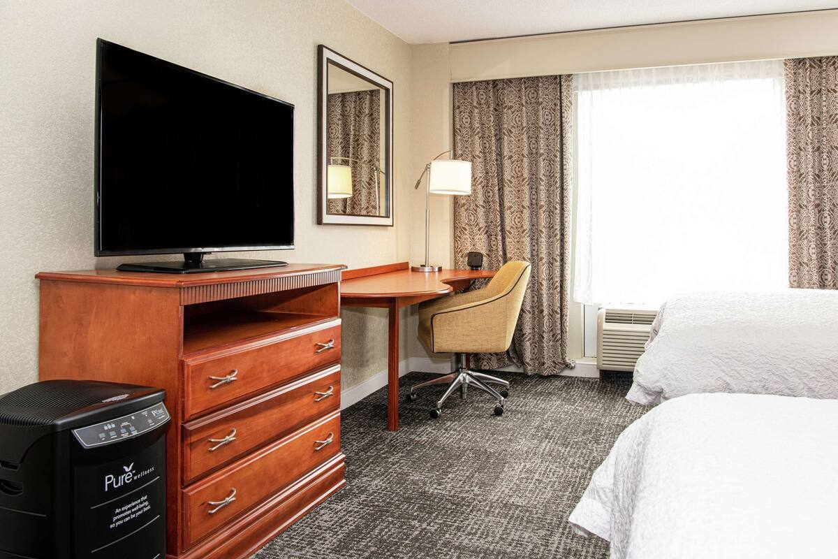 A hotel room with a bed, a desk with a chair, a television, a dresser, and a window with curtains.