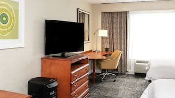 A hotel room with a bed, a desk with a chair, a television, a lamp, and a large window with curtains.