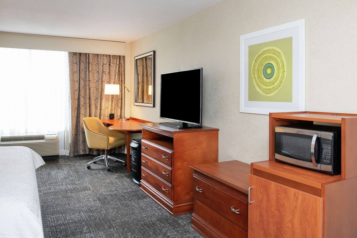 A hotel room with a bed, a desk with a television, a microwave, a chair, a dresser, and a framed artwork on the wall.