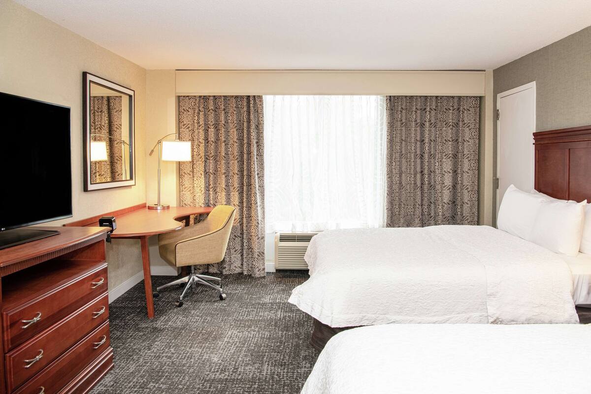 A hotel room with two beds, a desk, a chair, a TV, and a large window with curtains.