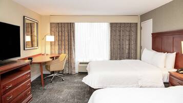 A hotel room with two beds, a desk, a chair, a TV, and a large window with curtains.
