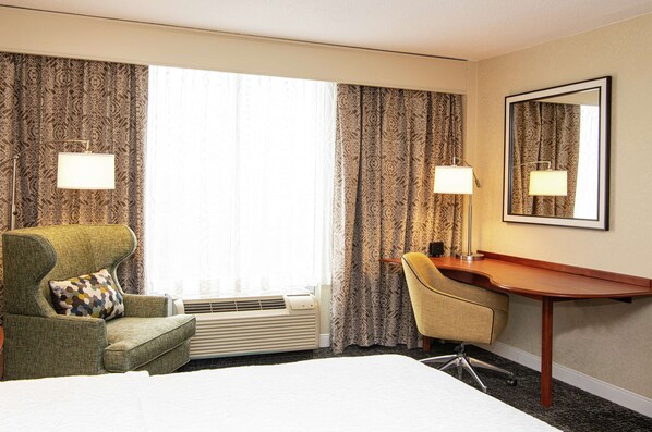 A hotel room with a bed, a desk, a chair, and a large window with patterned curtains. - Hampton Inn Parsippany (Parsippany)