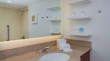 A bathroom with a sink, mirror, towel dispenser, and shelves with toiletries.