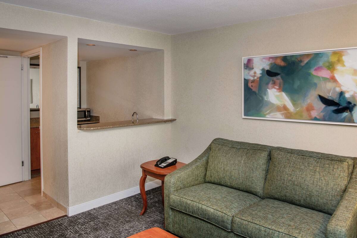 A living room with a green sofa, a small wooden side table with a telephone, a framed abstract painting, and a doorway leading to a kitchen.