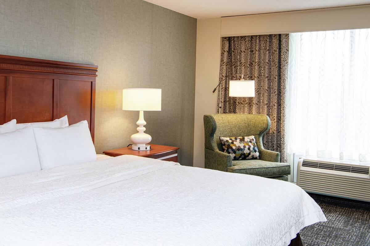 A hotel room with a large bed, a chair, a lamp, and a window with curtains.