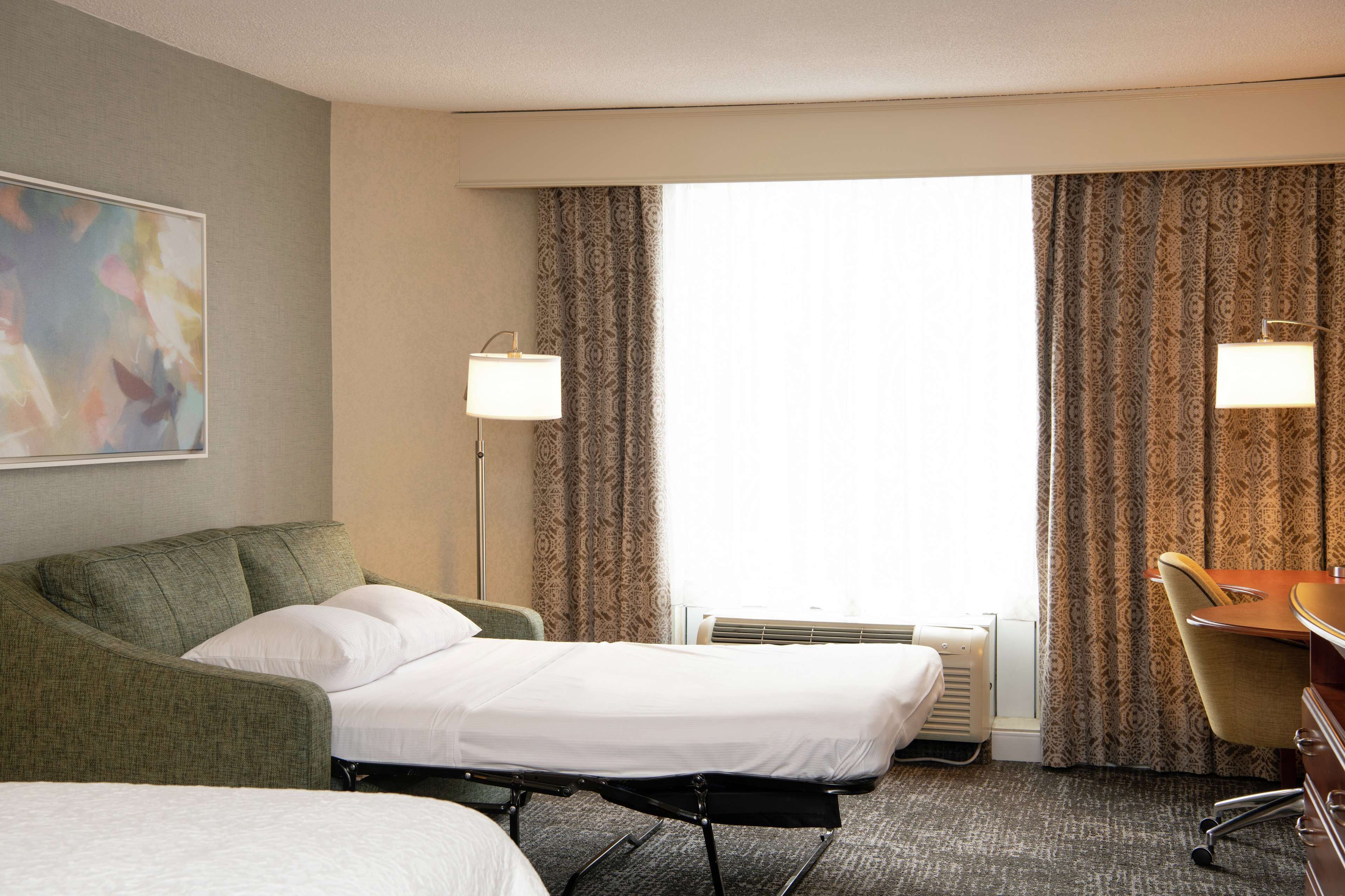 A hotel room with a bed, a desk, a chair, two lamps, and a painting on the wall.