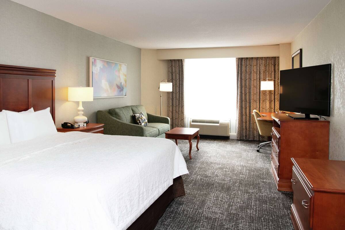 A hotel room with a large bed, a green armchair, a desk with a television, and a window with curtains.