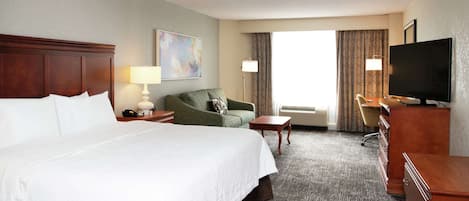 Habitación de hotel con una cama grande, un sillón verde, un escritorio con televisor y una ventana con cortinas.