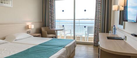 A hotel room with two beds, a balcony with a view of the sea, a small table and chair, and a desk with a mirror and lamp.