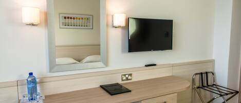 Habitación de hotel con tocador de madera, televisor de pantalla plana, espejo, cuadro floral enmarco y una botella de agua.