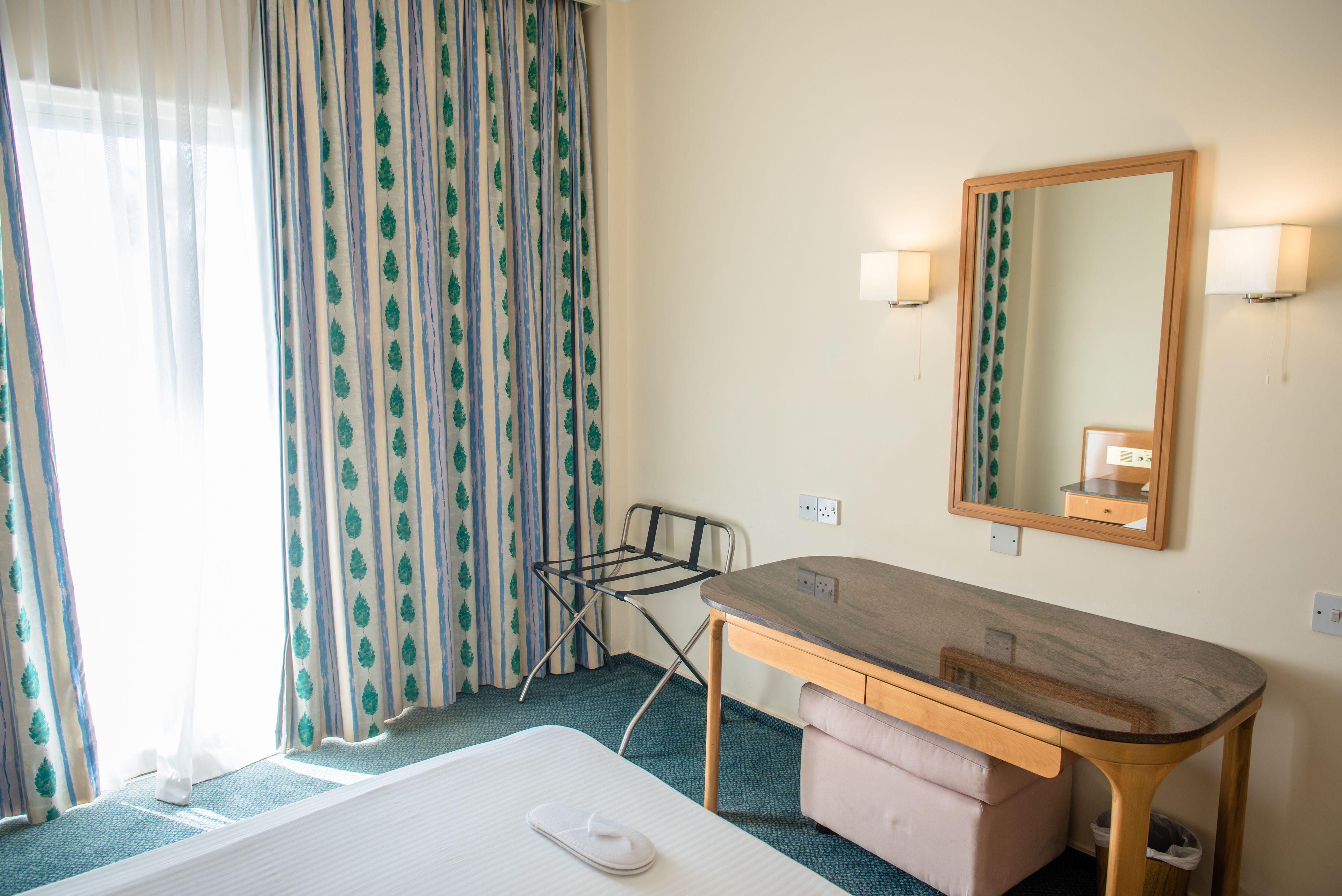 Habitación de hotel con cama, escritorio de madera, espejo y ventana con cortinas.