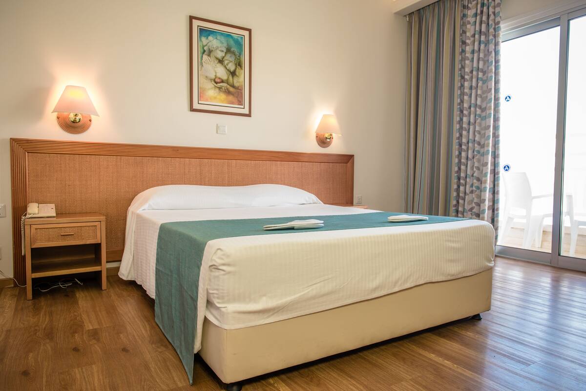 A hotel room with a large bed, wooden headboard, bedside lamps, a painting on the wall, and a view of the outdoors through a sliding door.