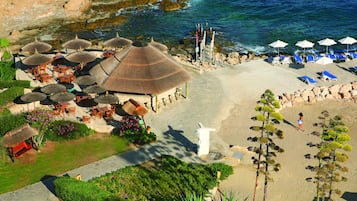 An aerial view of a beachside resort with thatched-roof umbrellas, lounge chairs, and a rocky coastline.