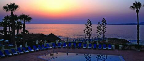 Zona junto a la piscina con sillones de descanso y sombrillas, con vista a un cuerpo de agua al atardecer.