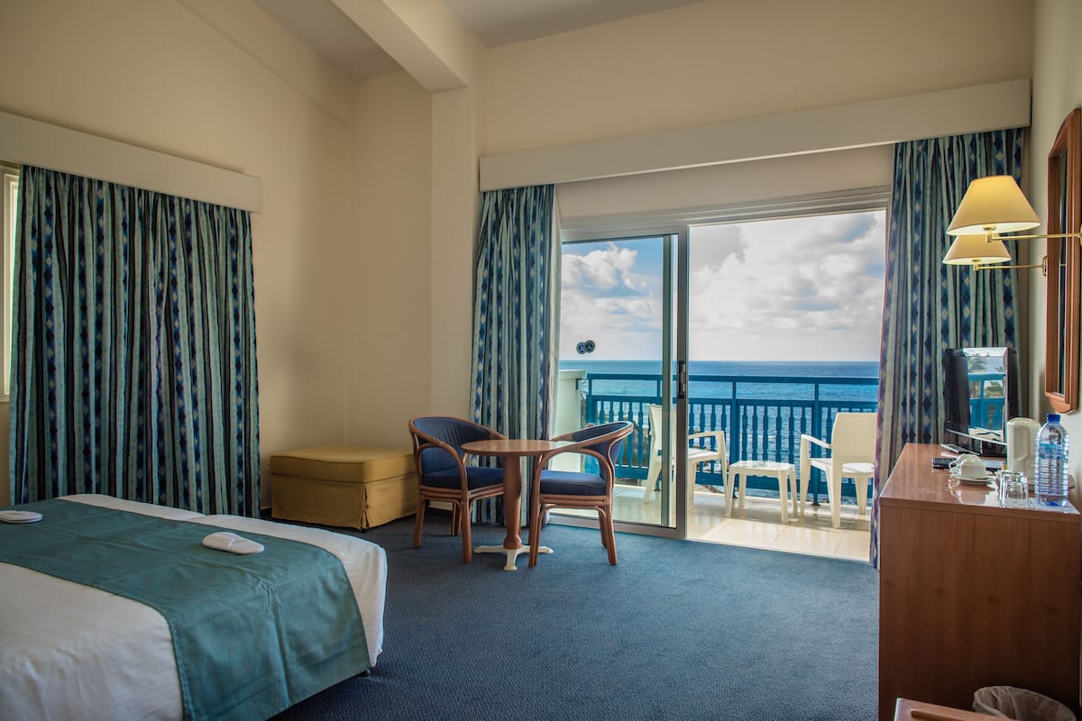 A hotel room with a bed, two chairs, a small table, a desk, and a view of the ocean through a sliding glass door.