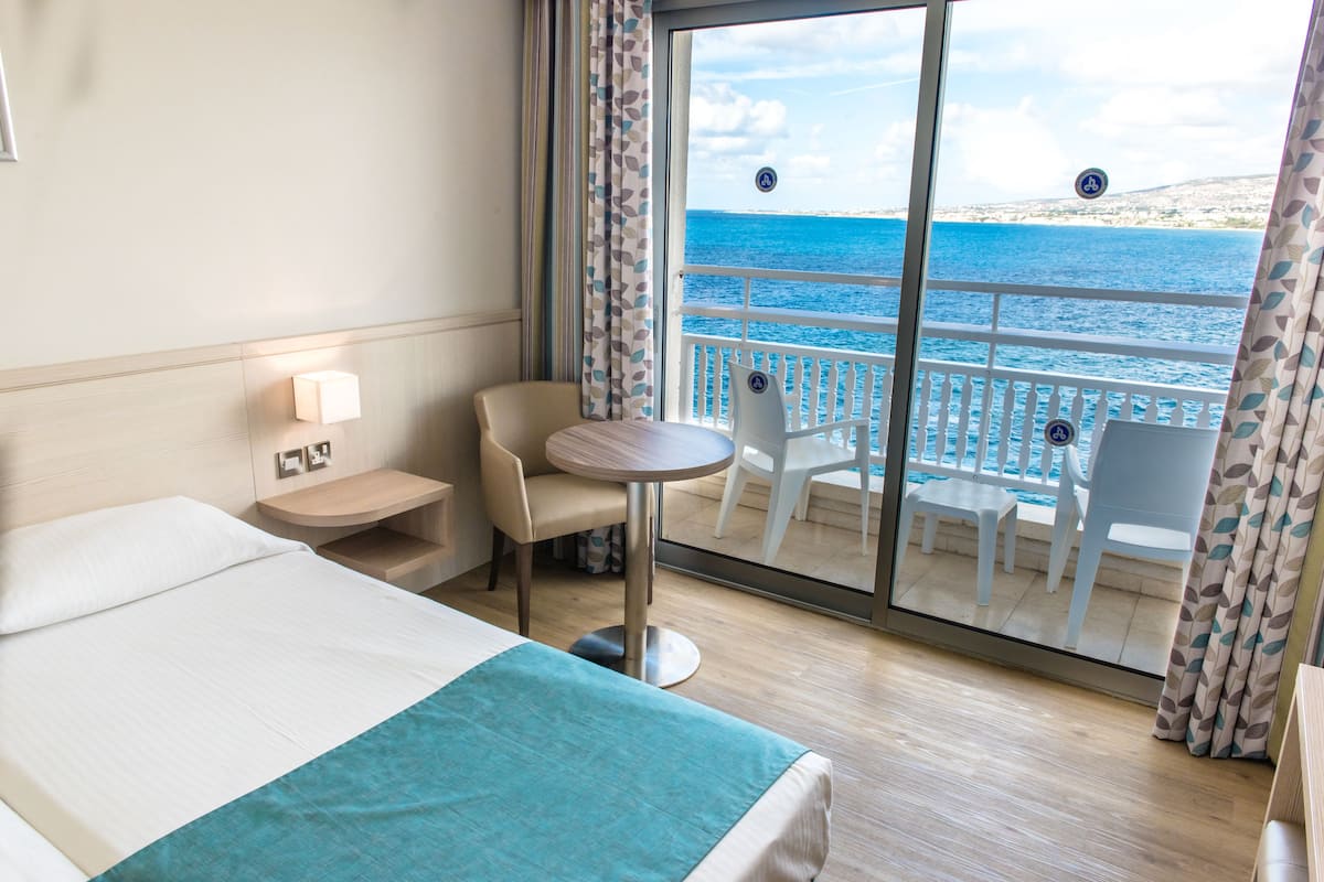 A hotel room with a bed, a small round table, a chair, and a balcony with a view of the sea.