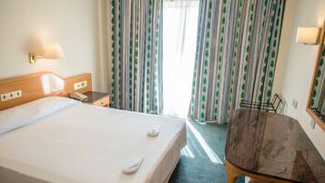 A hotel room with a bed, bedside table, lamp, and a small desk with a chair.