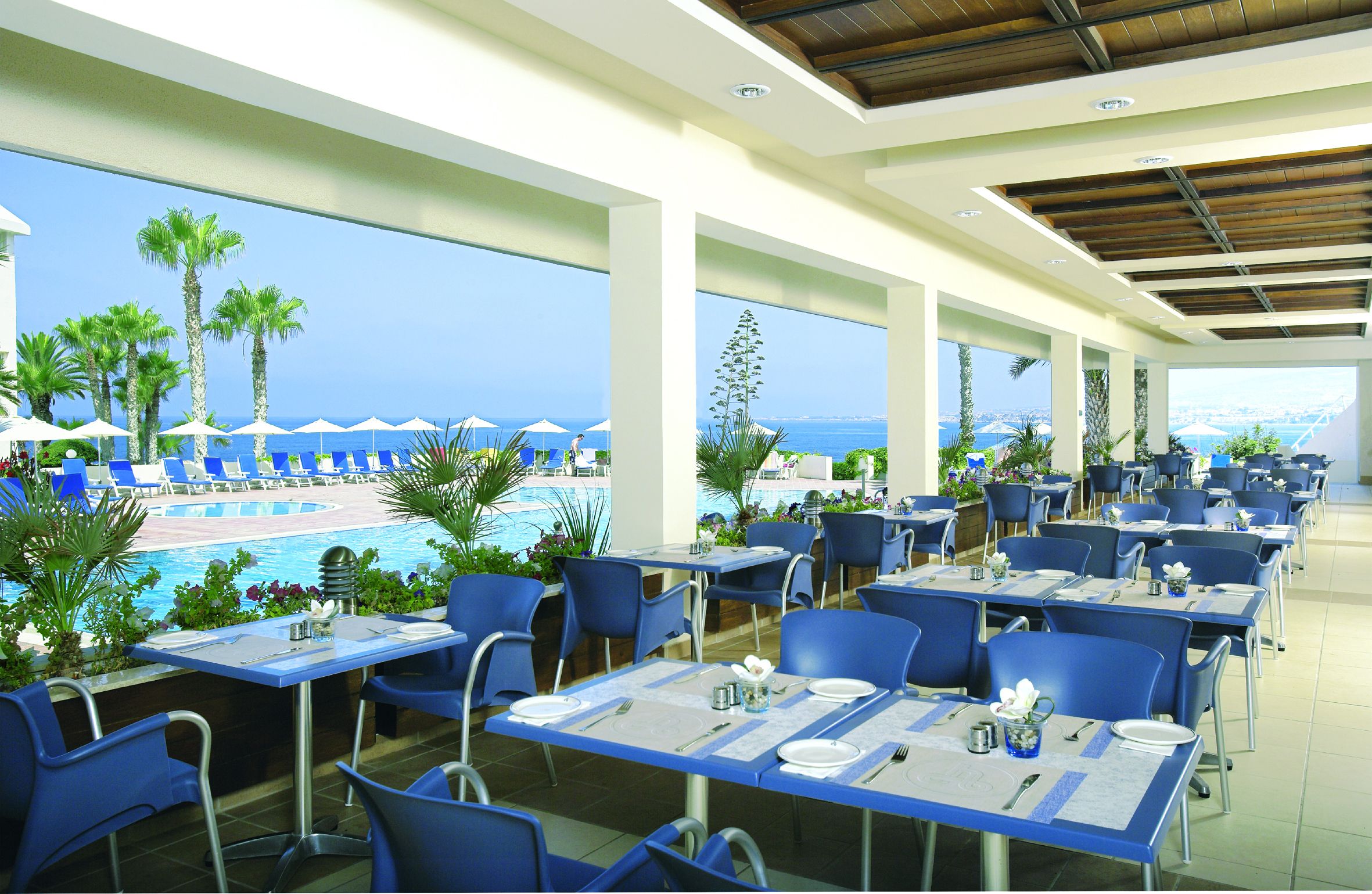 A restaurant with outdoor seating overlooking a pool and the ocean. Tables are set with blue chairs and white tablecloths.