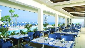 A restaurant with outdoor seating overlooking a pool and the ocean. Tables are set with blue chairs and white tablecloths.